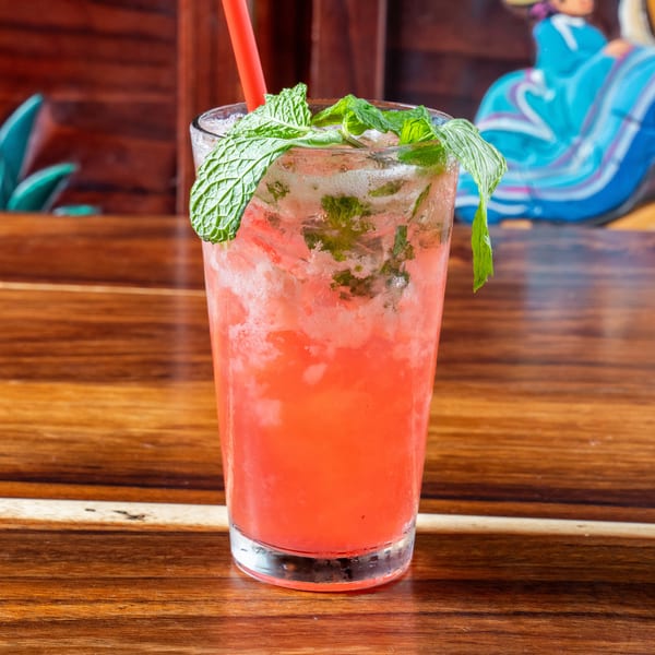 A vibrant pink cocktail with ice, fresh mint, and a red straw on a wooden table.