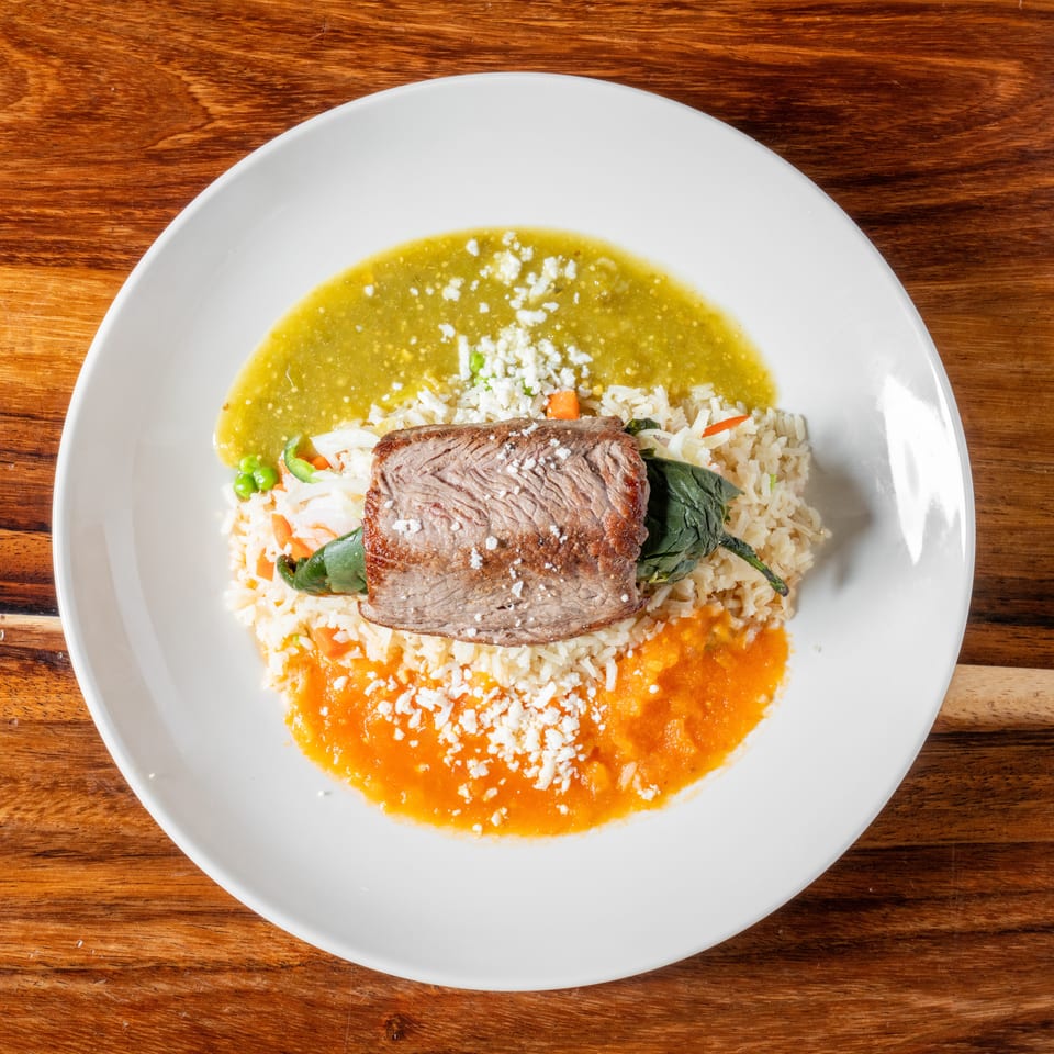 Steak with rice, green and red sauces, and crumbled cheese on a white plate.