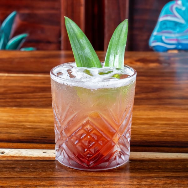 A vibrant red and orange cocktail with ice and green garnishes sits on a wooden table.
