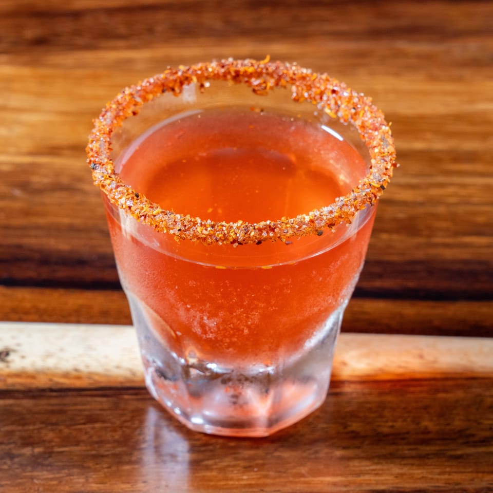 A vibrant red cocktail in a glass with a chili-salt rim, on a wooden table.