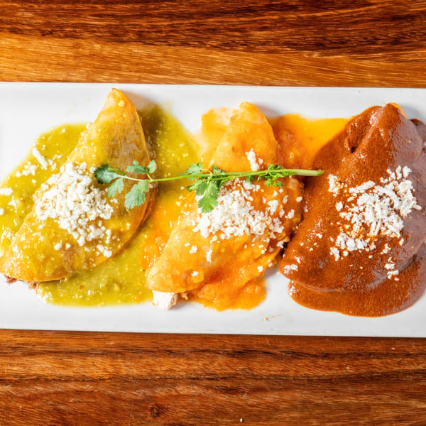 Three enchiladas with green, orange, and mole sauces, topped with cheese and cilantro on a white plate.