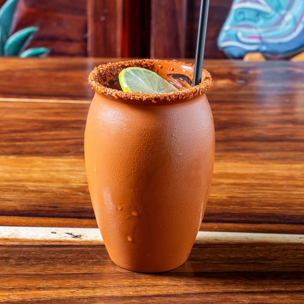 A chilled, terracotta-colored drink with a chili-lime rim, lime wedge, and black straw on a wooden table.