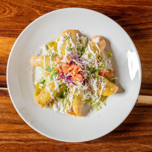 Taquitos topped with green sauce, sour cream, cheese, lettuce, tomatoes, and red cabbage.
