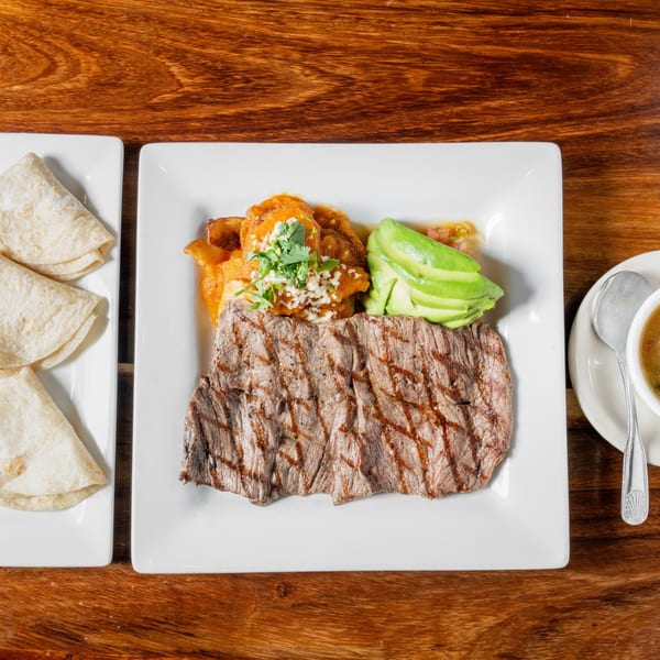 Grilled steak, avocado, and a side of tortillas and soup on a wooden table.