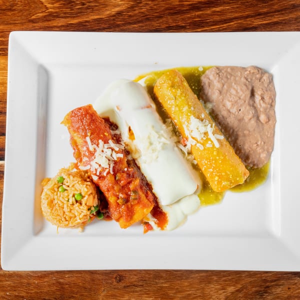 A plate of three tamales with red and green sauce, rice, and refried beans on a wooden table.