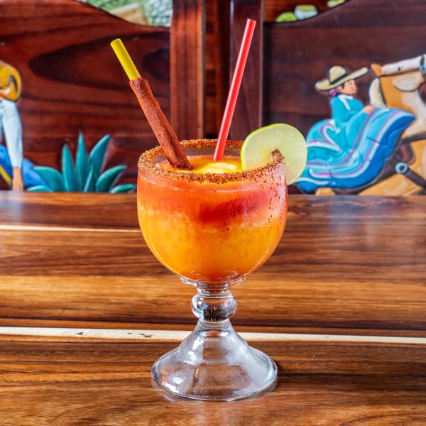 A vibrant orange drink with a chili-dusted straw and lime slice, on a wooden table with carved figures.