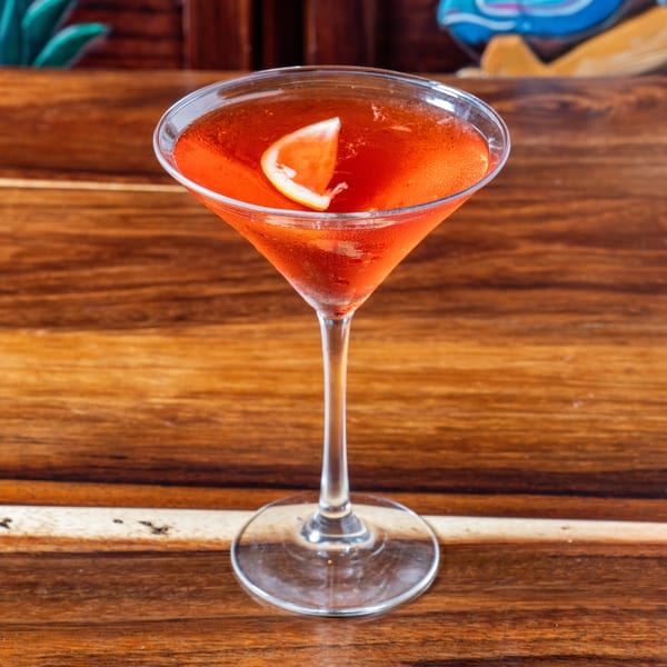 A vibrant red cocktail in a martini glass, garnished with a citrus wedge, on a wooden table.