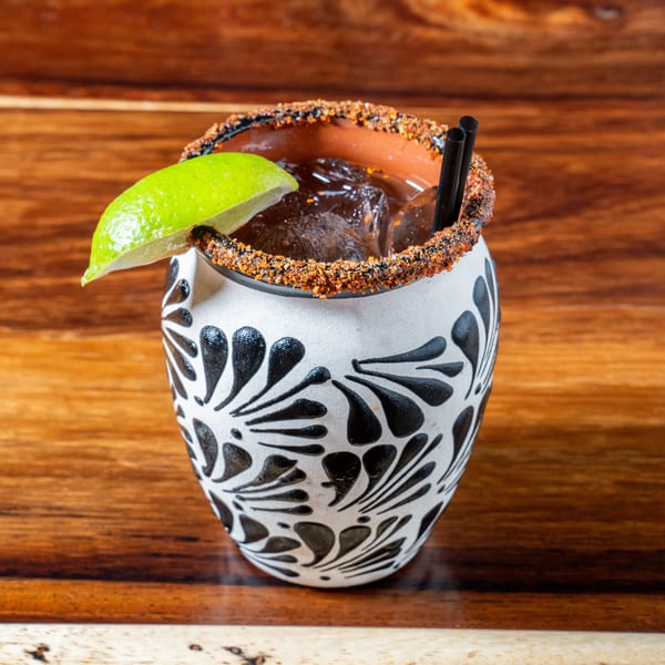 A dark cocktail with ice, a lime wedge, and two black straws in a decorative white and black mug.