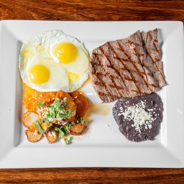 Grilled steak, two fried eggs, chilaquiles, and refried beans with cheese on a white plate.