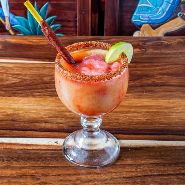 A vibrant red frozen drink in a goblet, rimmed with spices, garnished with lime and a chili straw.