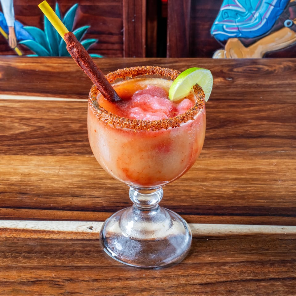 A vibrant red frozen drink in a goblet, rimmed with spices, garnished with lime and a chili straw.